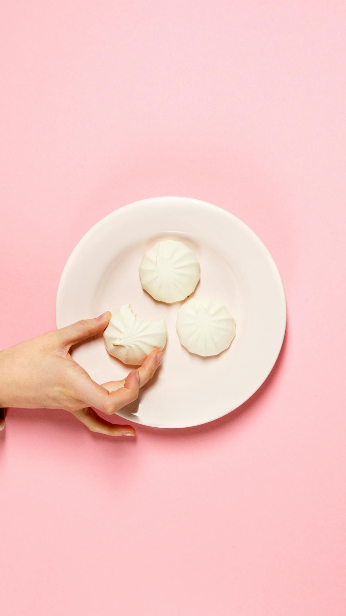 A hand reaching for white cookies on a plate against a vibrant pink background.